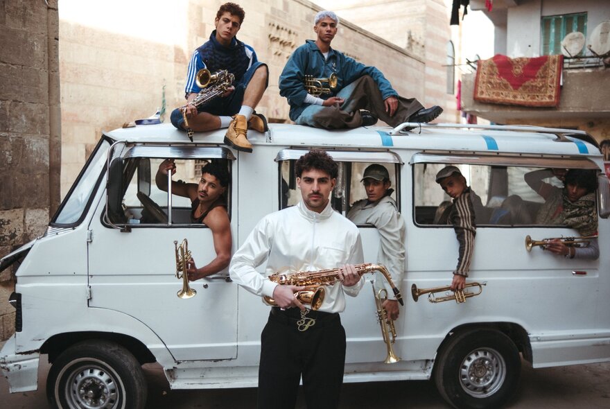 The musical artist Saint Levant, standing in front of a white van parked in a street and holding a saxophone, accompanied by a group of musicians seated inside the van or on top of it all holding trumpets and other musical instruments.