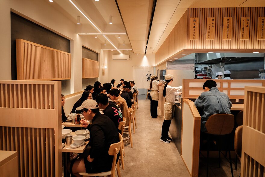The warm timber interior of Ginza Kagari restaurant in Melbourne, with patrons eating at tables and staff standing near the open kitchen.
