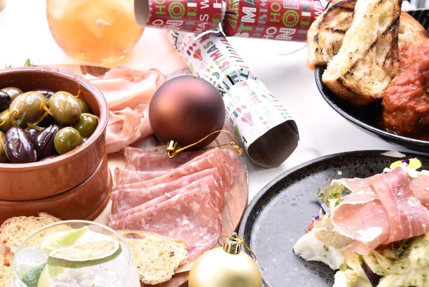 A table filled with plates of food including an antipasti platter, olives, bread and Christmas bon bon crackers.