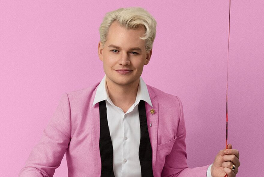 Comedian Joel Creasey in a pink suit with undone bowtie against an identically pink wall, holding the string of a balloon (unseen).