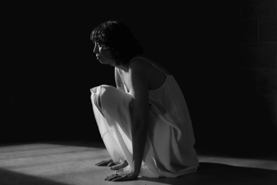 A black and white image of a crouching woman in a white camisole nightie, in a beam of light surrounded by darkness.