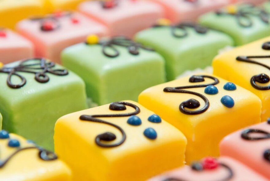 Small cube-shaped brightly coloured desserts, decorated with piped chocolate.