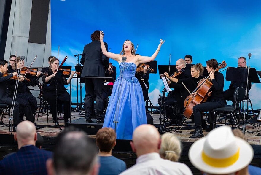 An opera singer in a blue dress in front of a small orchestra, the audience in the foreground. 