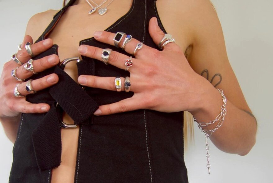 Model wearing a black waistcoat, holding her hands covered in rings over her chest.