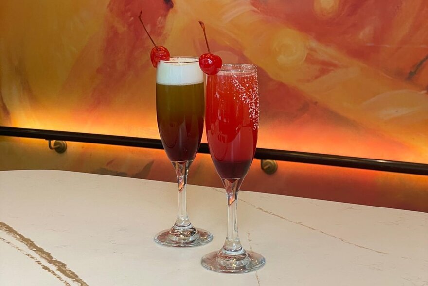 Two cocktails in flute glasses, with glace cherry garnishes, on a marble table top inside Miss Mi restaurant.