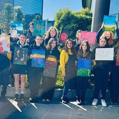 A row of smiling teenagers standing outside a building, all holding up artworks they've created. 