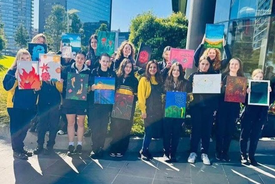 A row of smiling teenagers standing outside a building, all holding up artworks they've created. 