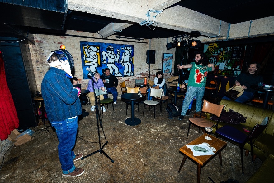 People in a basement space, some sitting and watching a person performing in front of a microphone.