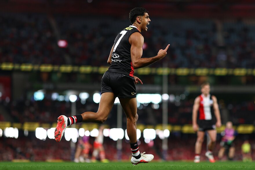 A St Kilda AFL player running and celebrating on the grassy field, with his finger pointing in front of him, blurred players and spectators in the background.