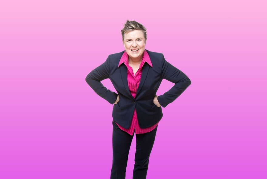 Comedian Monica Dullard wearing a dark pink shirt and black suit jacket and pants, posing with her hands on her hips against a hot pink background.