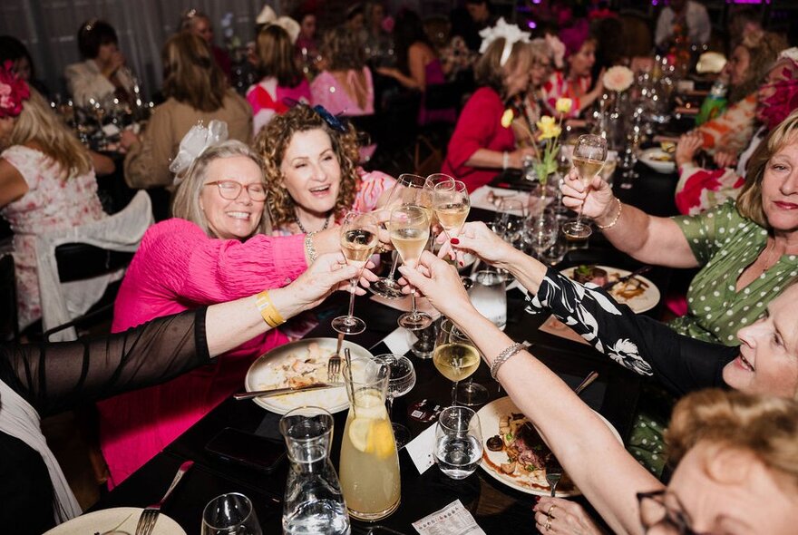 A group of women in spring racing finery clinking glasses over a restaurant table.
