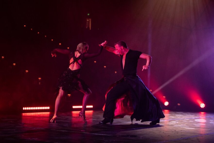 Two performers posed in a flamenco-like dance move, on a dark stage, with red back lights.