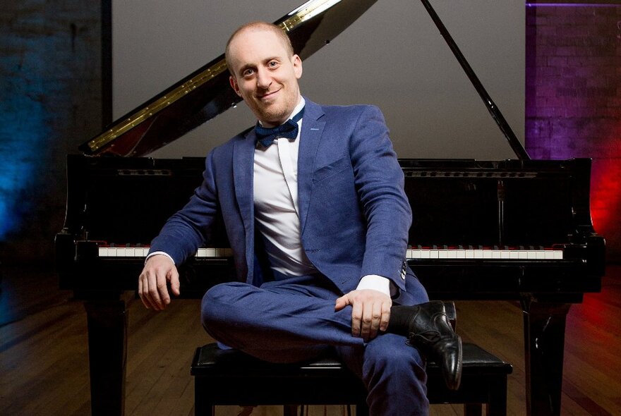 Australian pianist, Simon Tedeschi, sitting on a piano stool in front of a grand piano, wearing a blue suit.