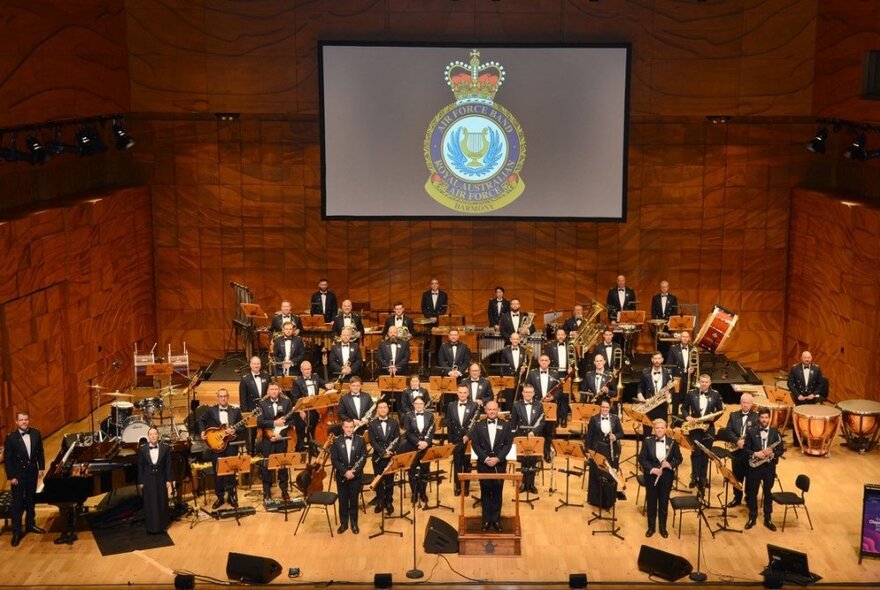 Looking down in an auditorium at a the RAAF band on a stage, members all standing, with a large tv screen and the RAAF logo.