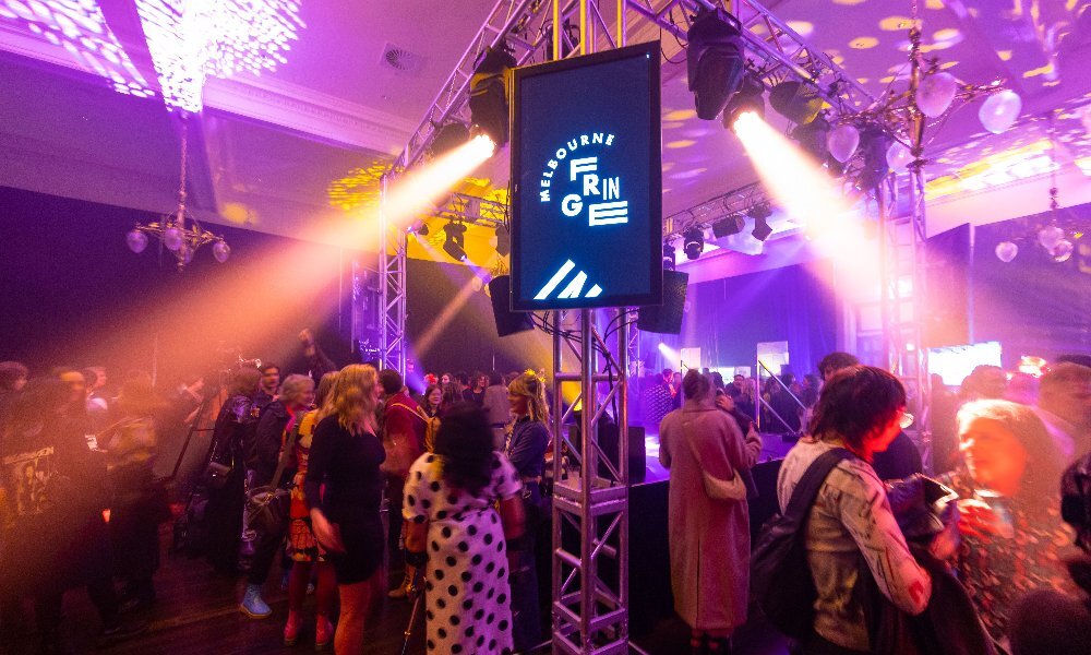 A crowd of people gathered around a Melbourne Fringe Festival stage with purple and yellow lights.