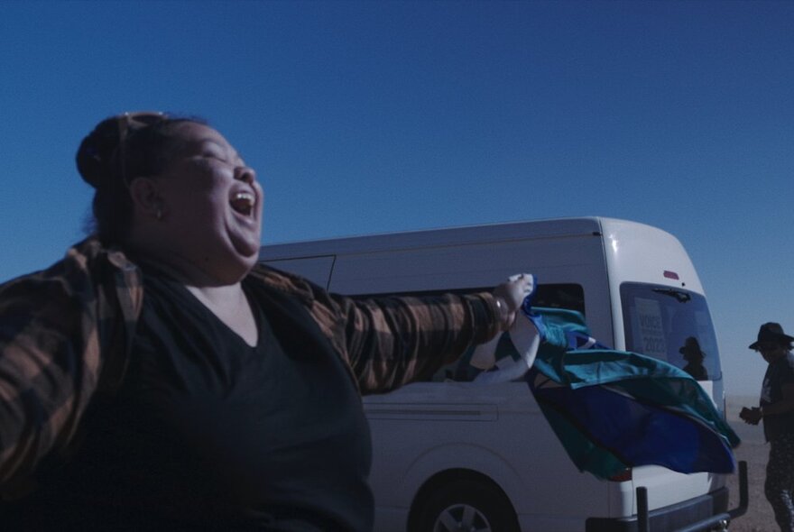 A woman standing in front of a white passenger van with her arms widespread with a look of pure joy on her face.