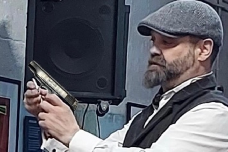 Bearded man wearing a cap and waistcoat holding a gun in front of a speaker.