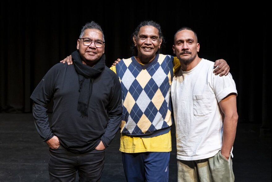 Three men standing arm in arm, the central man wearing an Argyle vest and yellow T-shirt.