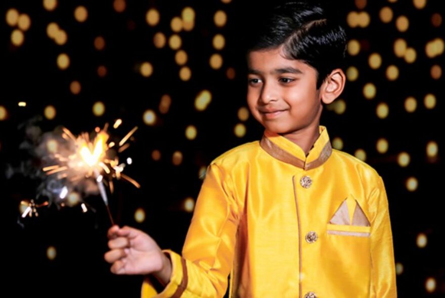 An Indian boy wearing a yellow satin coat, holding a sparkler in front of a sparkling black background.