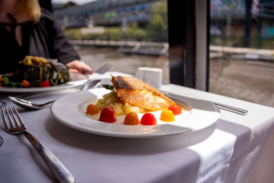 A  table on a floating river cruise boat with plates of food  and cutlery, with views of the Yarra River in the background.