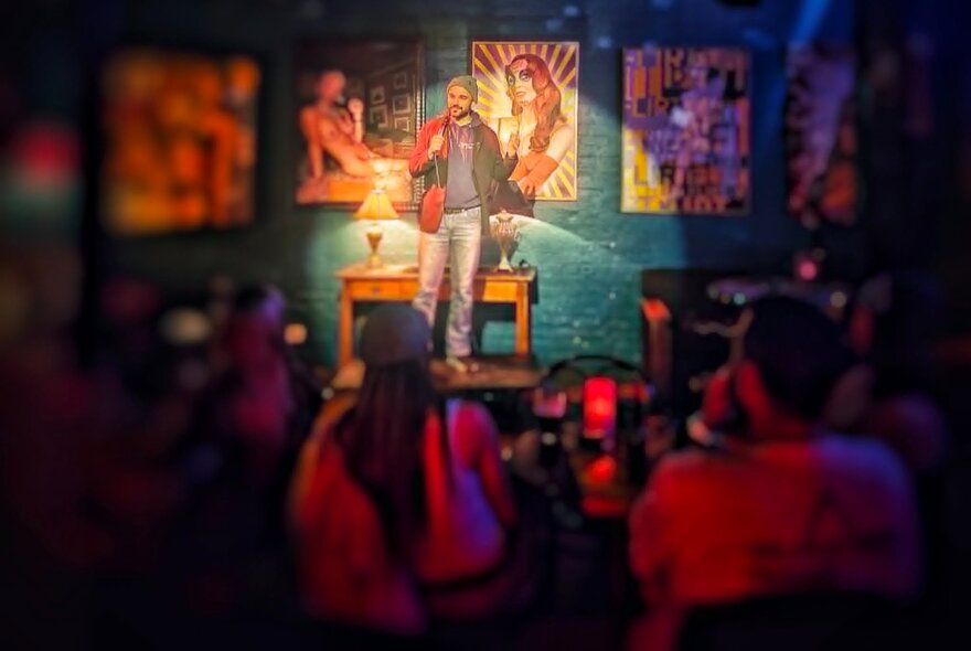 A person holding a microphone performing stand-up comedy on a small stage at Fad Gallery Bar, in front of a live audience.