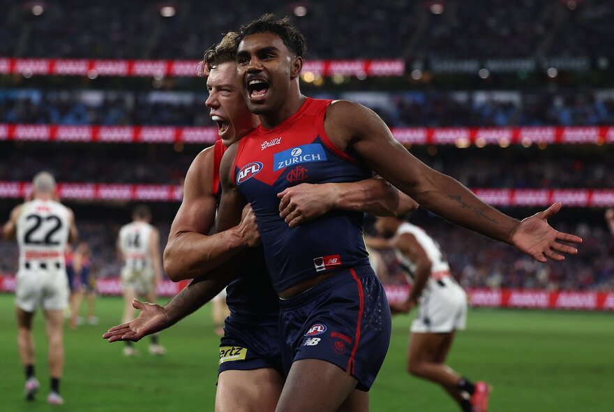 A Melbourne player hugs another Melbourne player from behind in celebration on the football field. 