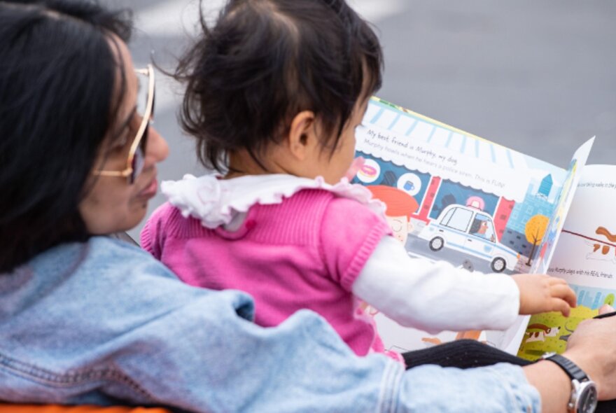 A toddler sitting on their mother's lap, both of them looking at a picture book, in the open space of a library.