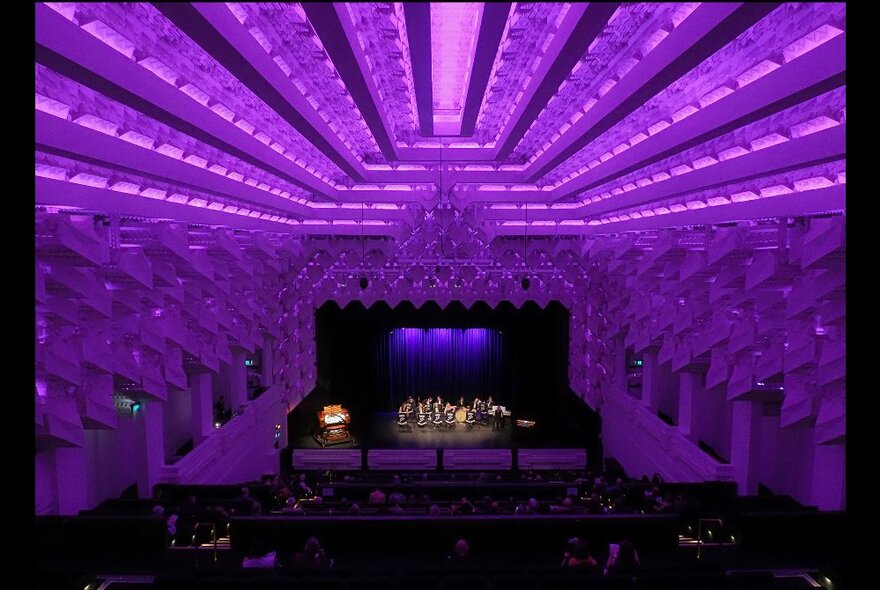 Interior of the Capitol Theatre, awash with purple lights that show the architectural details of the ceiling and walls, a band playing on the stage in the far distance.