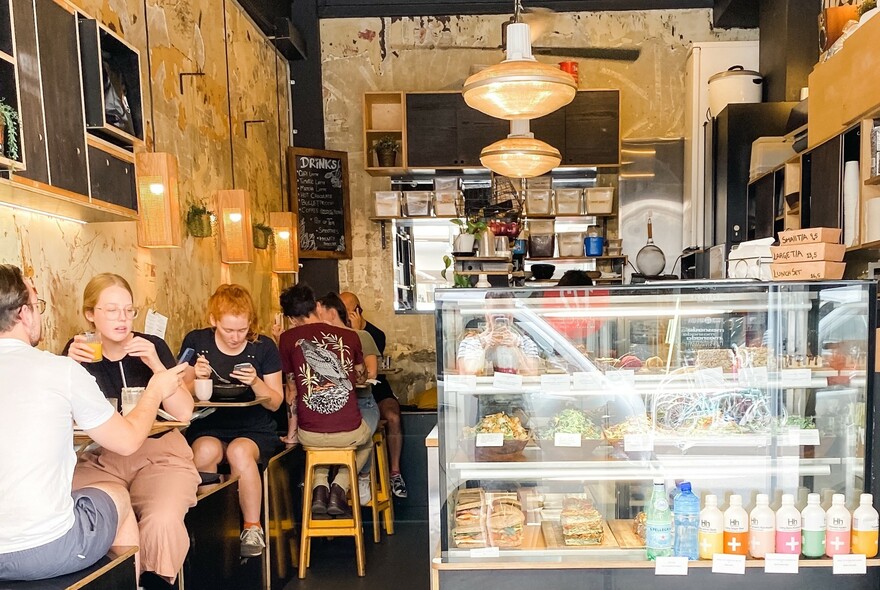 Cafe interior with people at tables, glass-fronted food counter and bottles of drinks.