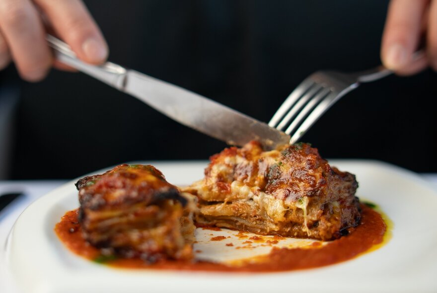 A person cutting into a serve of lasagne with a knife and fork.