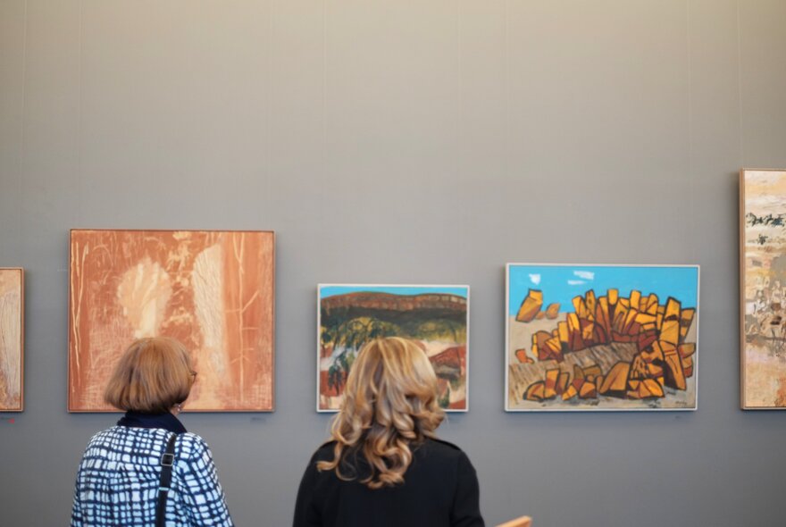 Artworks of Australian landscapes hanging on a grey wall with the back of two women looking at them.
