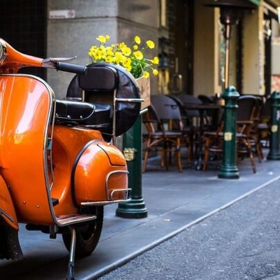 An orange Vespa parked on a sidewalk in a city laneway with cafe tables and chairs in the background.