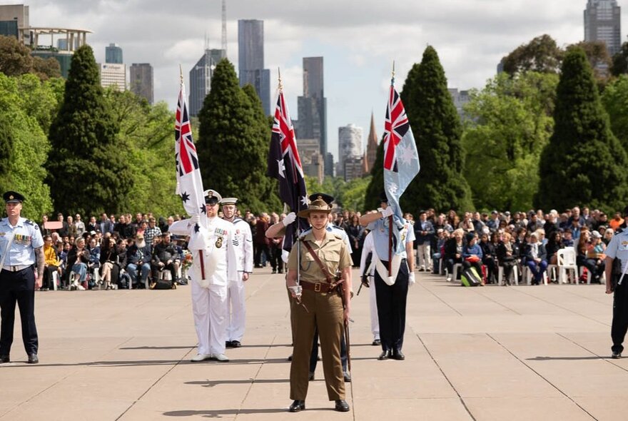 Service people standing on the forecourt of the Shrine with city buildings and massed crowds in the background.