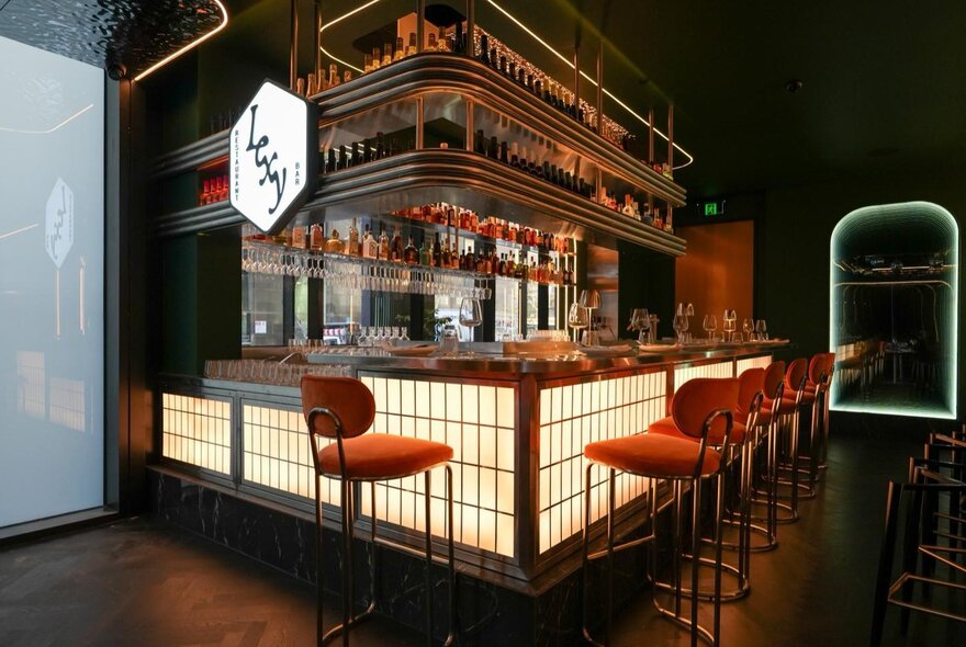 Restaurant's bar area with Asian-inspired decor, illuminated screen detail, red stools and wooden shelving.