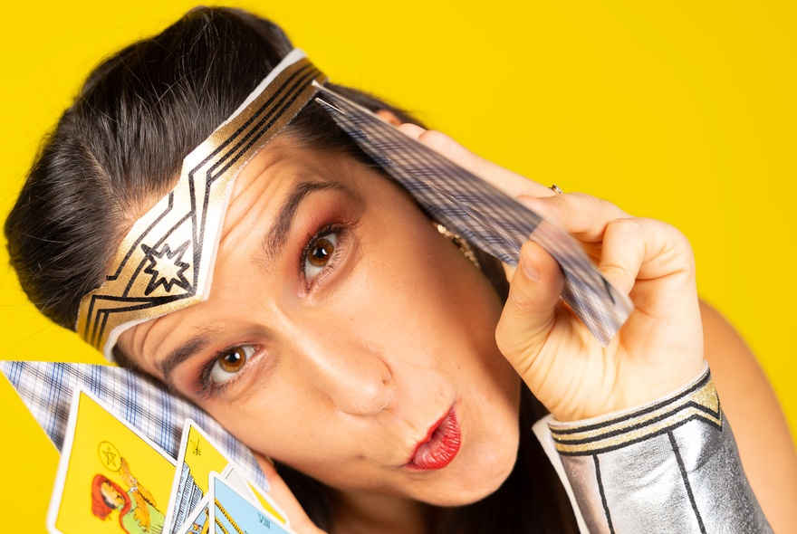 Close-up of a woman's face dressed in a Wonder Woman costume, featuring a silver tiara and silver cuffs, looking directly at the camera, holding tarot cards with a surprised expression.
