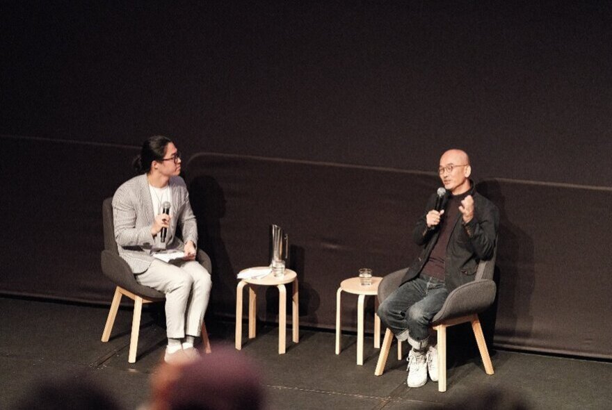 Two people sitting on a black stage, having a conversation.