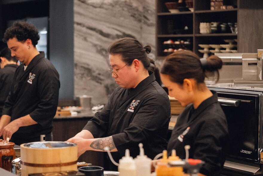 Three Japanese chefs in black preparing food at a Japanese restaurant, a dumpling steamer and sauce bottles in the foreground.