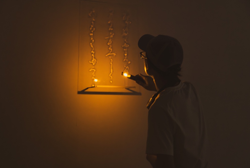 A person holding a small lit torch, interacting with a light-based art installation hanging on a wall in a dimly lit gallery space.
