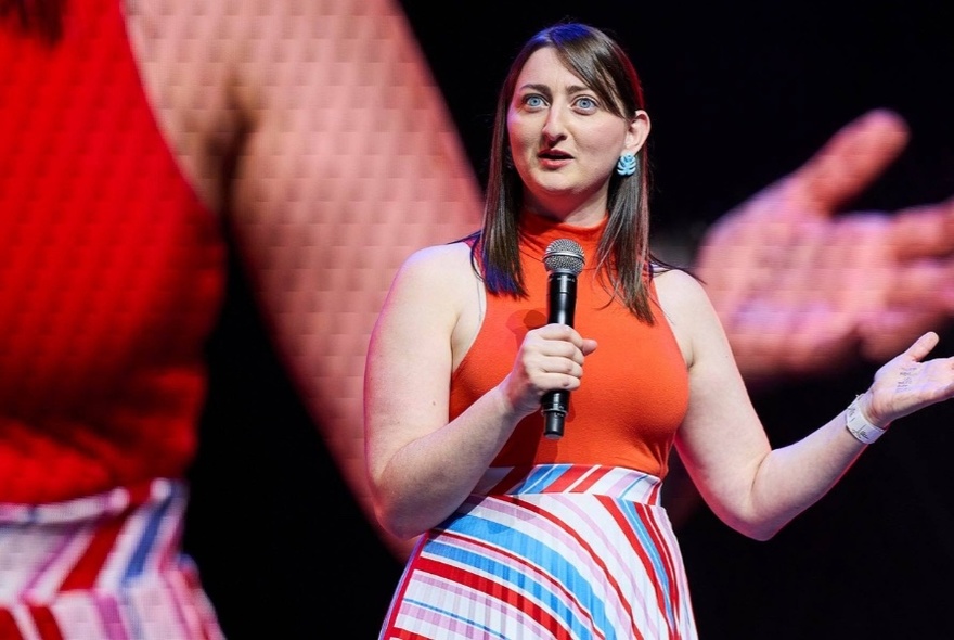 Comedian Sonia Di Iorio performing holding a microphone in front of large screen image of herself.