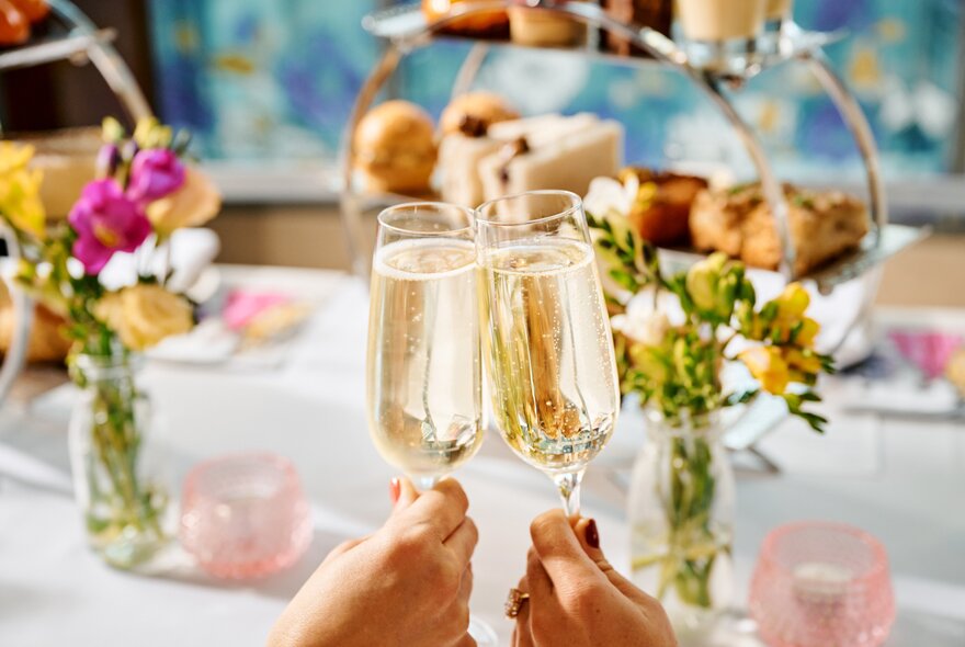 Two champagne glasses being held together, in front of a cake stand and floral table arrangements.
