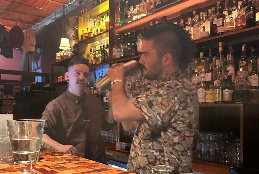 A person shaking a cocktail behind a bar while being instructed by a bartender.