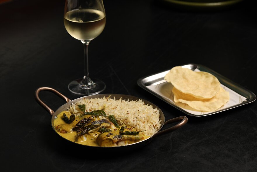 A dark table with a bowl of curry and a side plate of papadums, a glass of white wine at the back. 