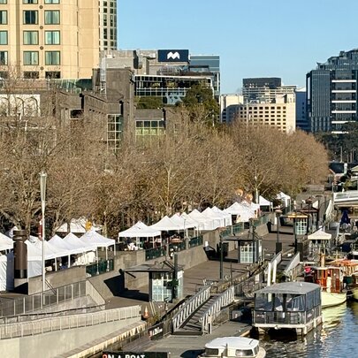 Aerial view of an outdoor market on Southbank promenade, showing the Yarra River with boats on the right, market umbrellas and stalls on the concourse and city buildings in the background.