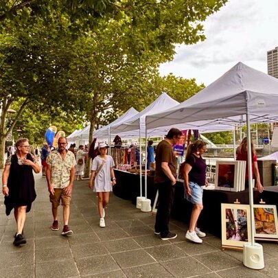 People walking past market stalls under white gazebos at an outdoor market, leafy trees on the left.