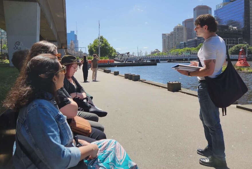 Tour leader talking to a group of seated walking tour participants near the banks of the Yarra River with city buildings visible in the background.
