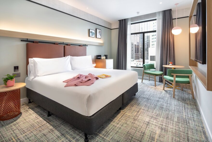 A hotel room with king size bed, white linen, green chairs near a small desk, and soft grey curtains.