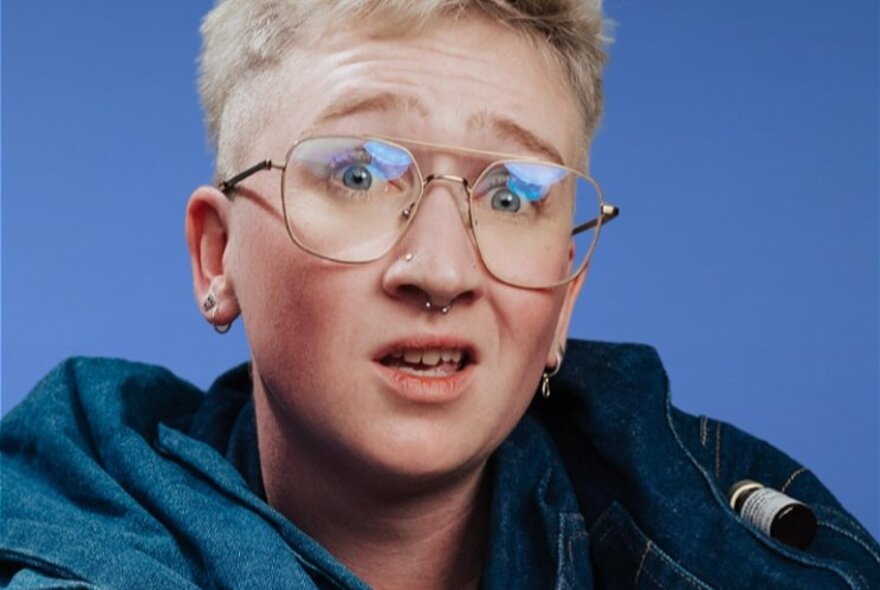 A person with short blonde hair, large gold-framed glasses and a septum ring, with a puzzled expression.