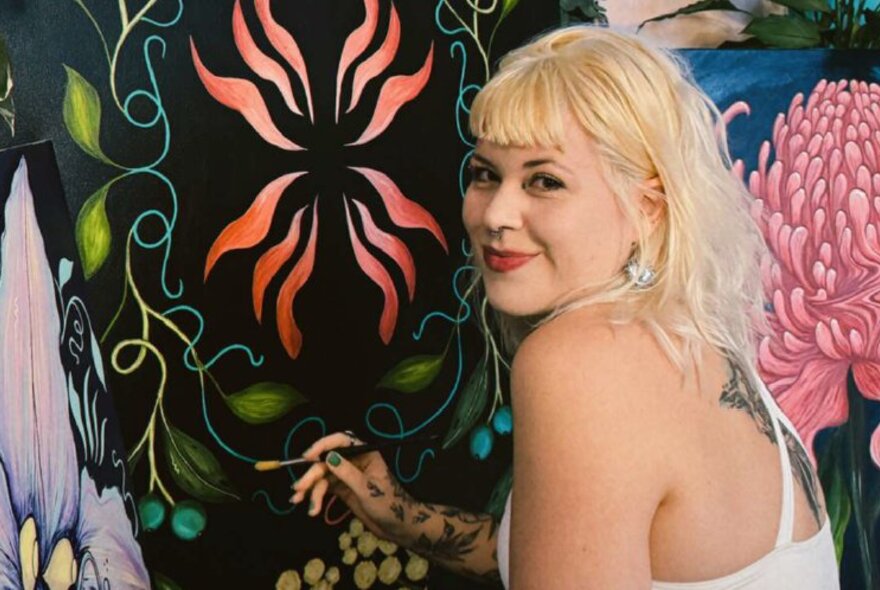 Blonde-haired artist applying paint to a black and floral artwork.