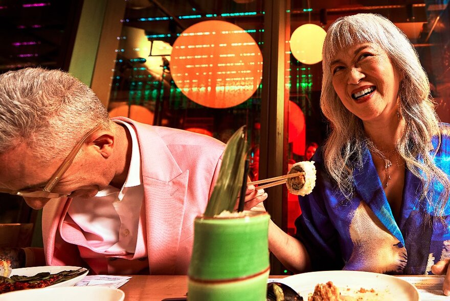 People enjoying a meal at an Asian restaurant, smiling woman using chopsticks, man wearing pink jacket laughing.