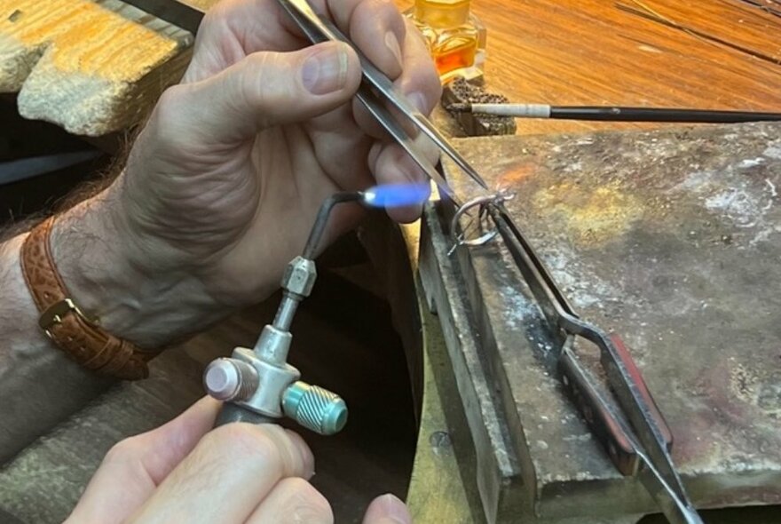 Hands holding a soldering iron and tweasers over a ring clamped in place on a workbench.